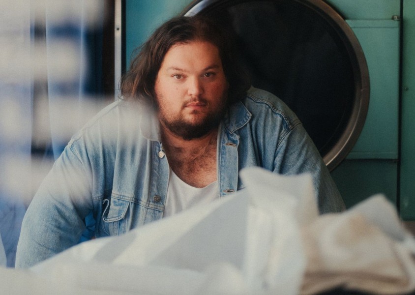 A man with dark hair and a beard wearing a white tshirt and denim shirtx
